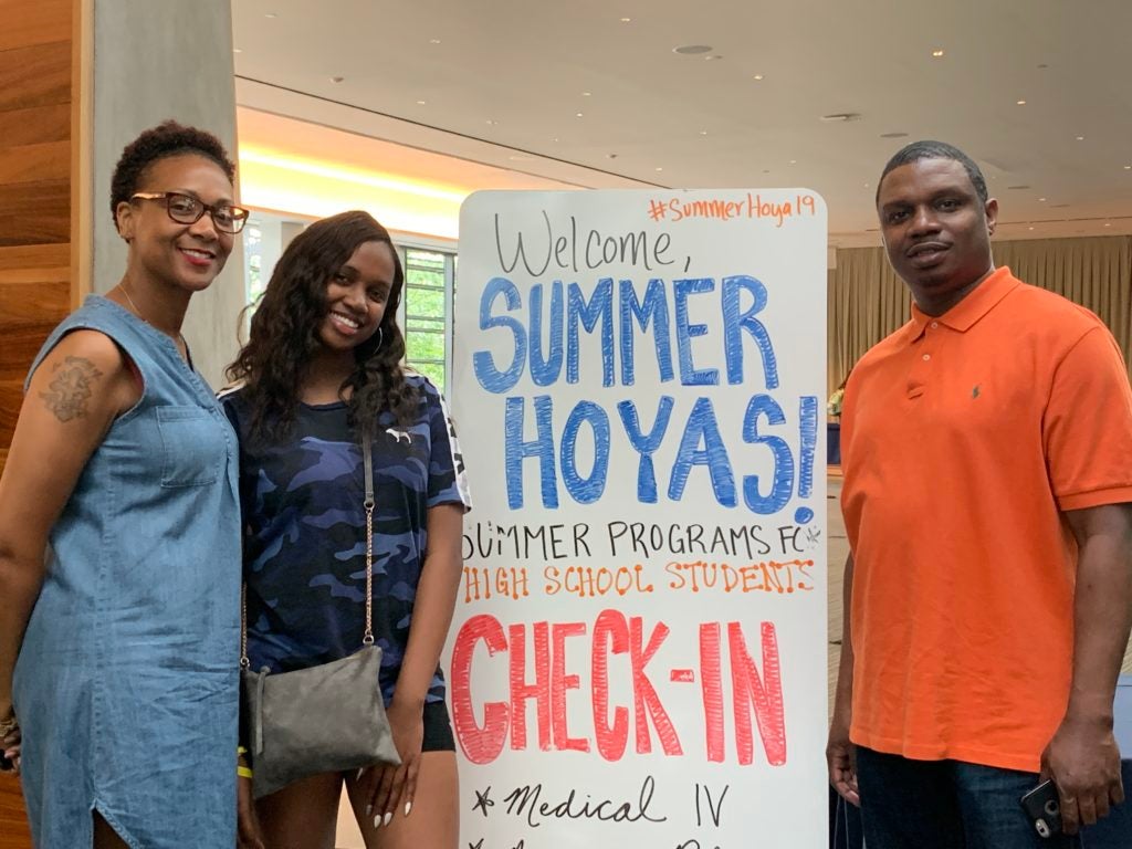 Two high school students and an adult stand near a sign that reads Welcome Summer Hoyas