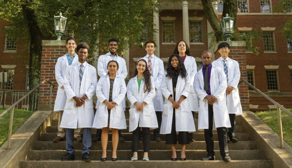 A group of students wearing white coats stands on the steps of the Med-Dent Building front entrance