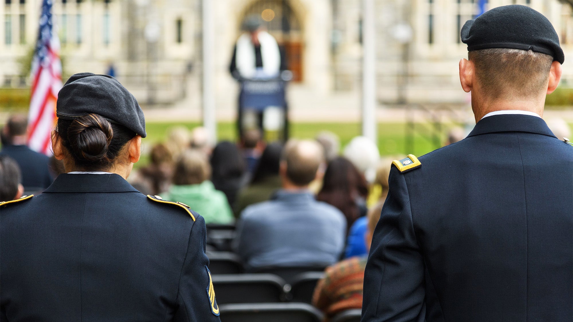 Two students in military uniform stand with their backs to the camera while in front of them a ceremony is taking place