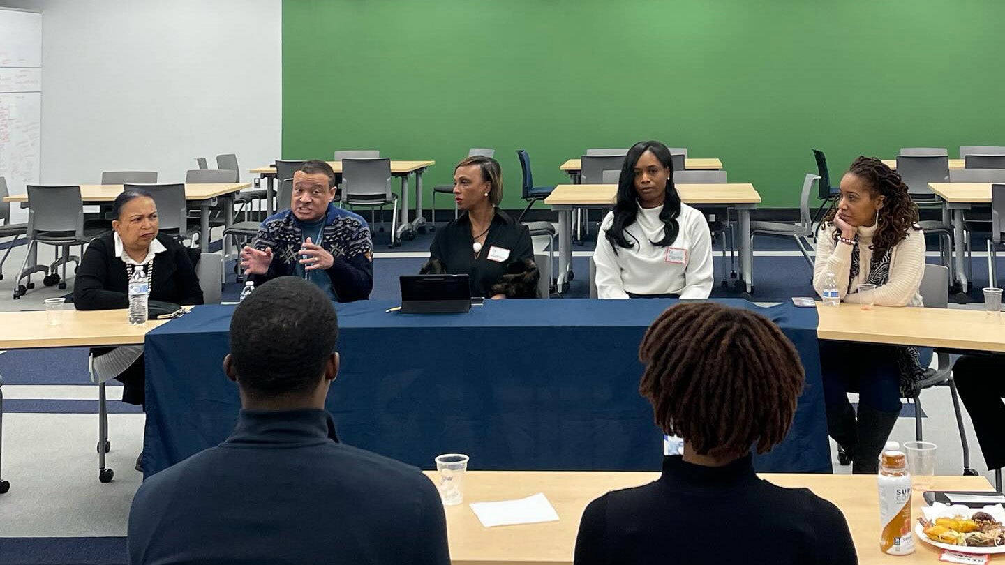 Five panelists sit at a table at the front of a room speaking to an audience