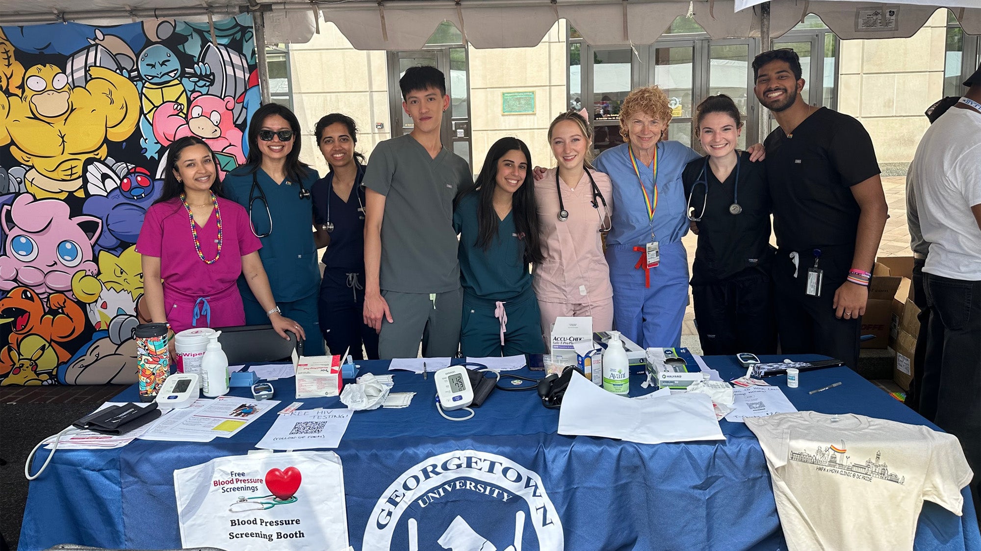 A group of volunteers stands side by side behind a table filled with materials for Pride attendees