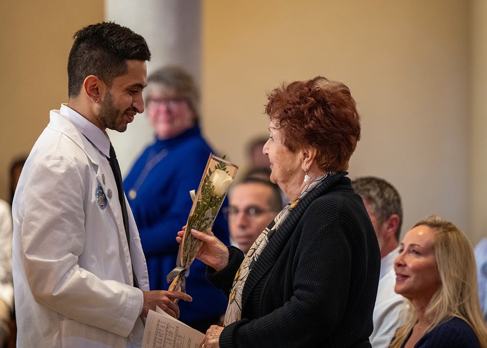 A medical student wearing a white coat hands a white rose wrapped in a plastic sleeve with a ribbon to a participant in the Donor Mass ceremony