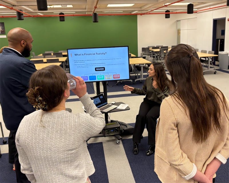 Nadia Samaha gestures toward a monitor on which her presentation is displayed as several people look on