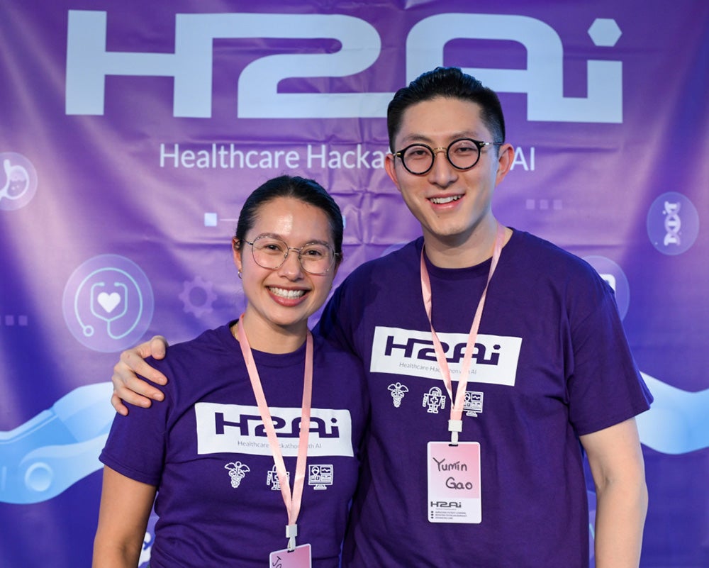 Jacqueline Sandling and Yumin Gao, both wearing purple H2AI T shirts, stand side by side in front of the H2AI banner