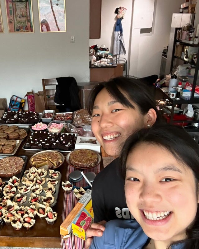 The Li sisters pose with their baked goods arrayed on a table behind them and the rest of their apartment in the background