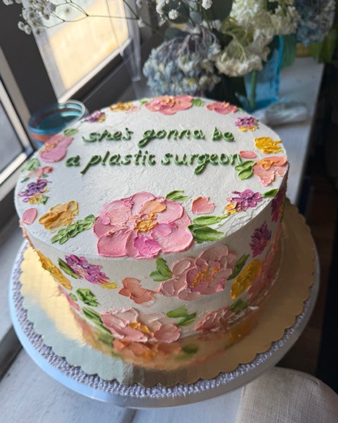 A lovely layer cake decorated with pink, yellow and violet flowers and the words she's gonna be a plastic surgeon in frosting