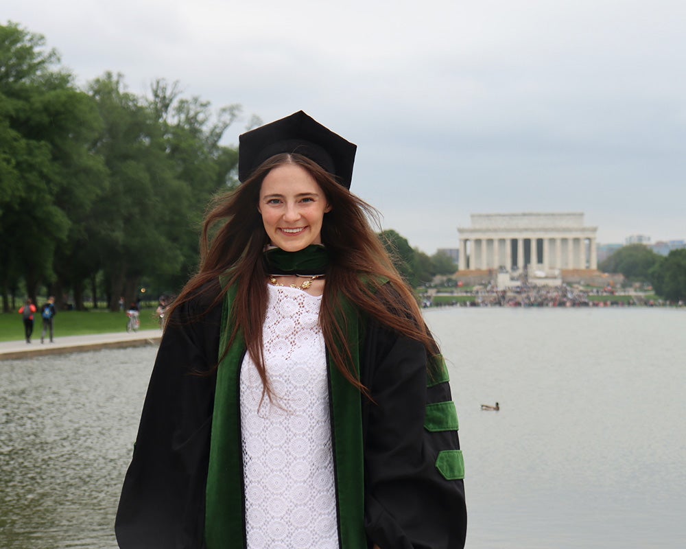 Sarah Lucas, wearing her graduation regalia, stands at the reflecting pool with the Lincoln Memorial in the background