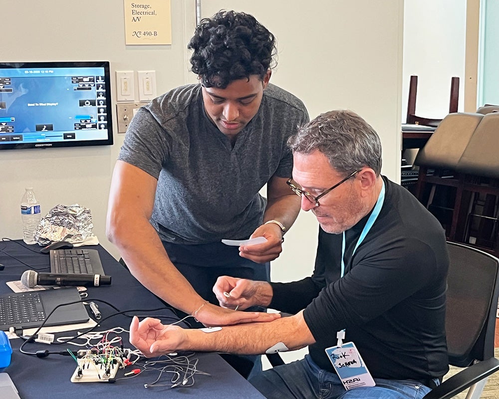 A medical student affixes an electrode to Richard Schena's inner arm, as wires poke from a circuit board on the table in front of him