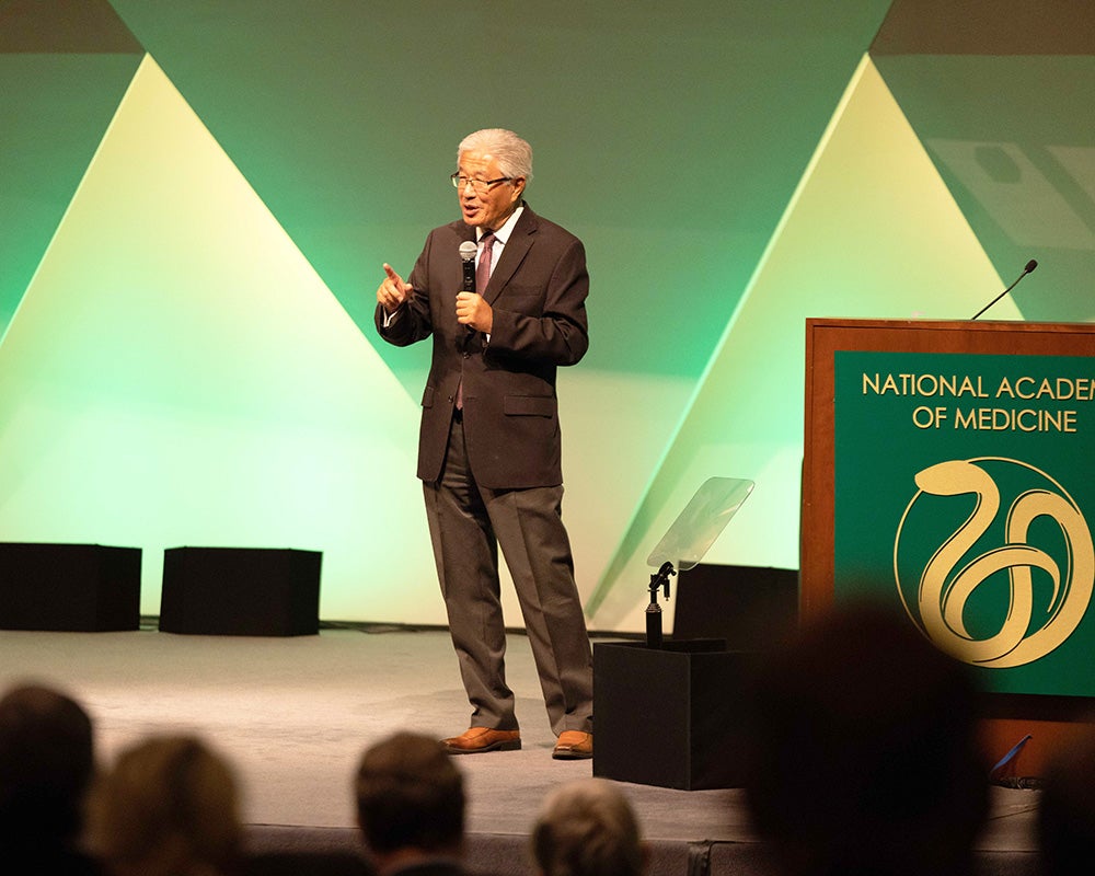 Dr. Dzau stands next to a podium with the National Academy's logo on it holding a microphone and speaking before an audience