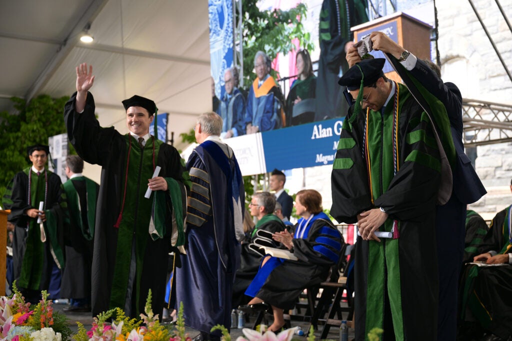 Two students onstage, one walking across with arm raised the other with head bowed to receive a hood