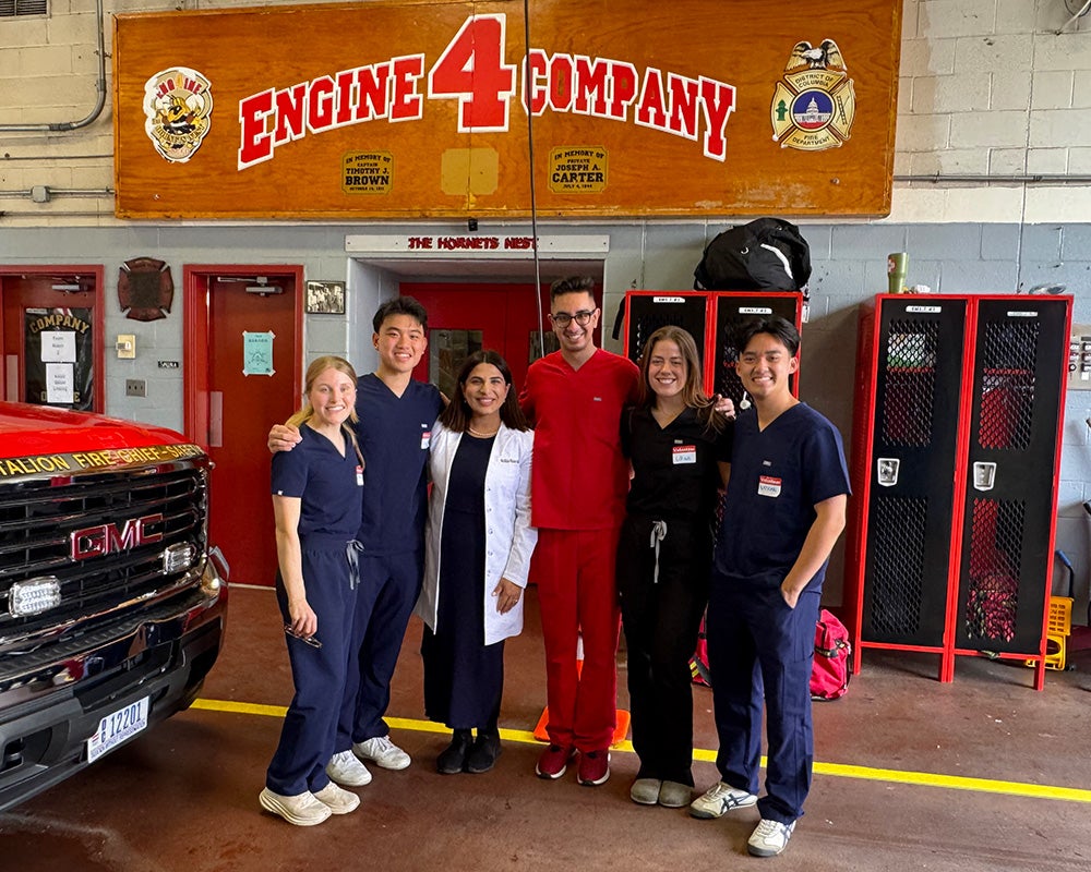 Six people stand together in a firehouse in front of a sign that reads Engine 4 Company