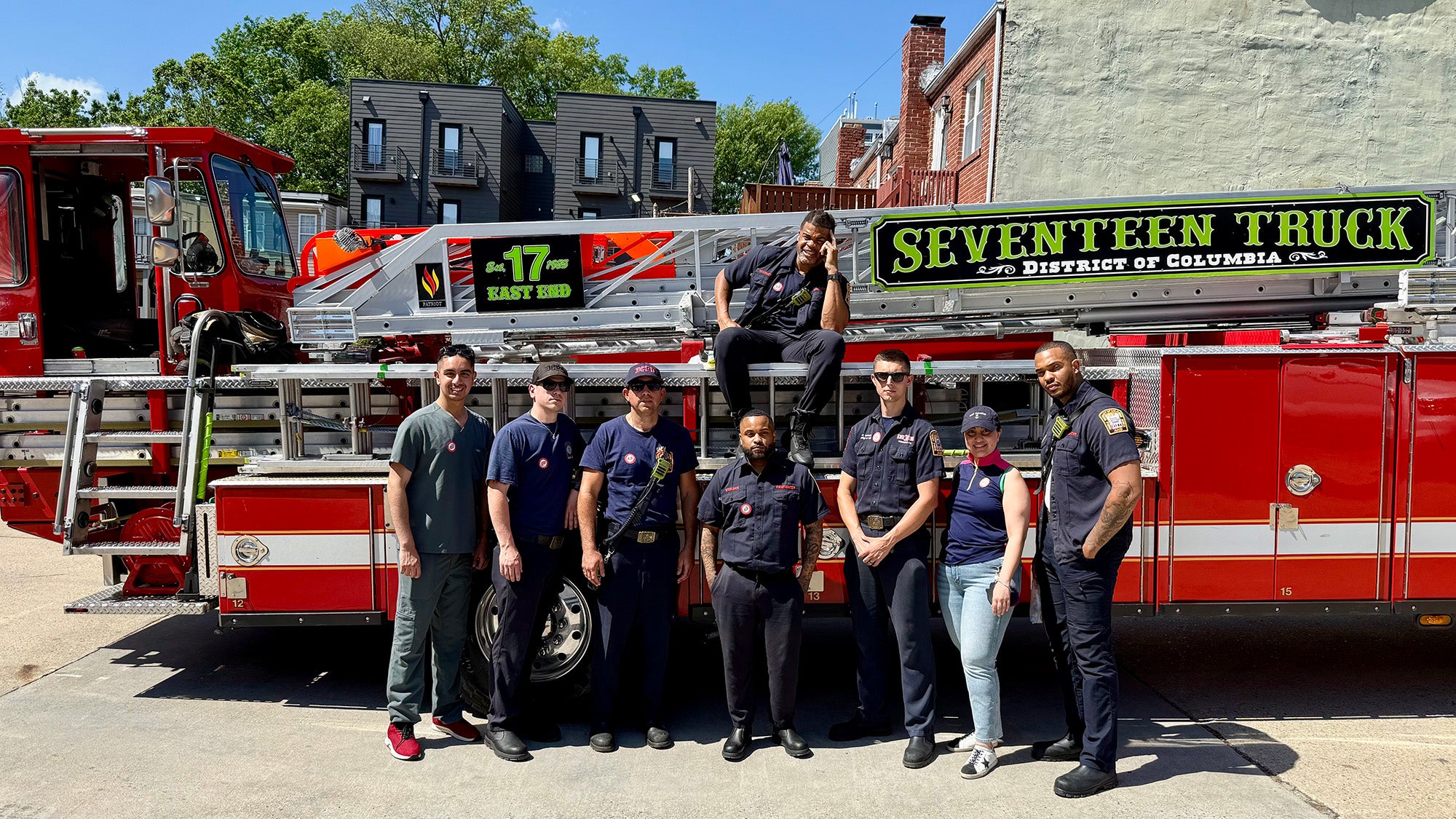 Seven people stand in front of a fire truck while one person sits on the truck behind them