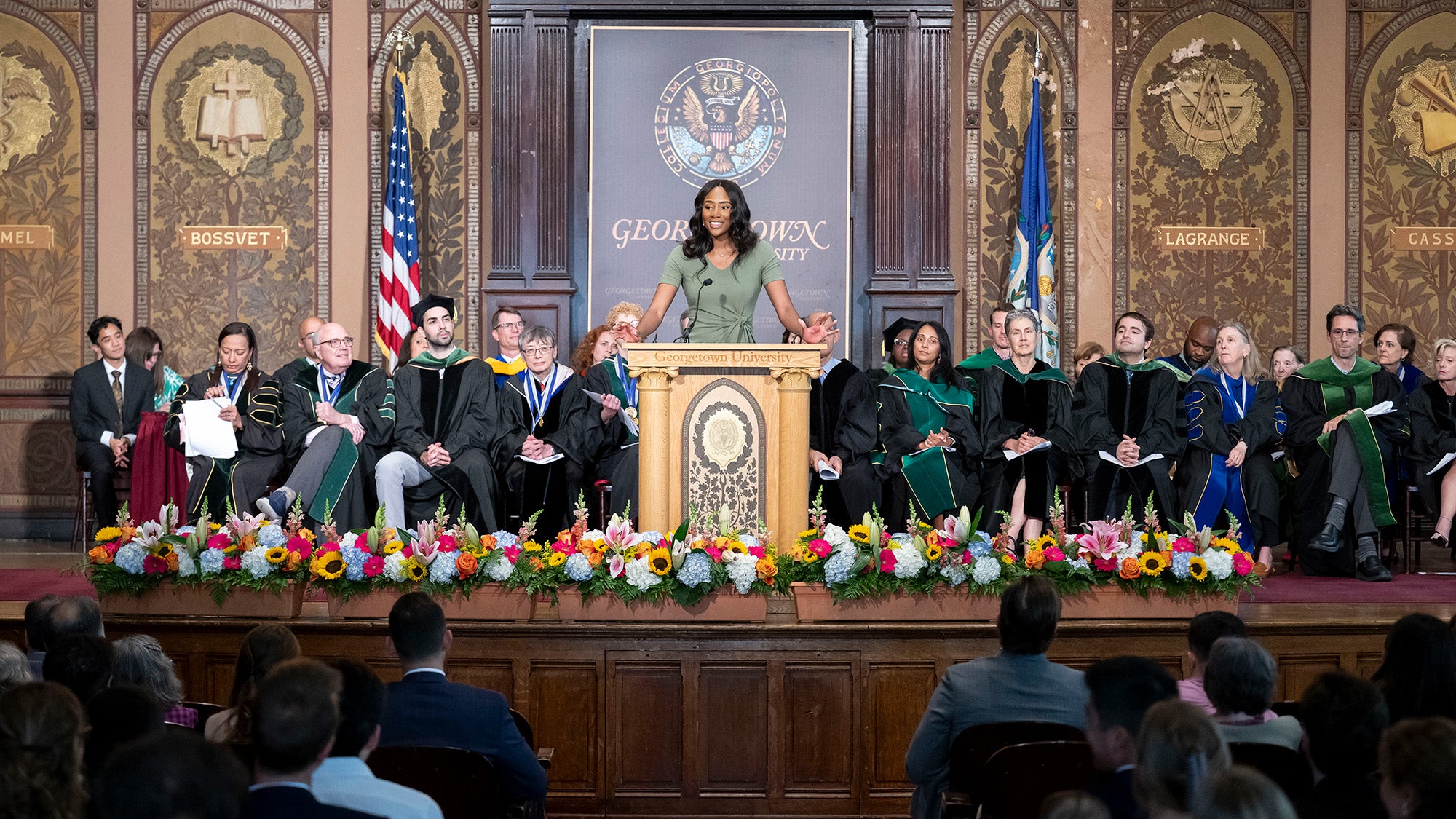 Tami Alade stands at a podium with the medical school faculty seated in rows behind her