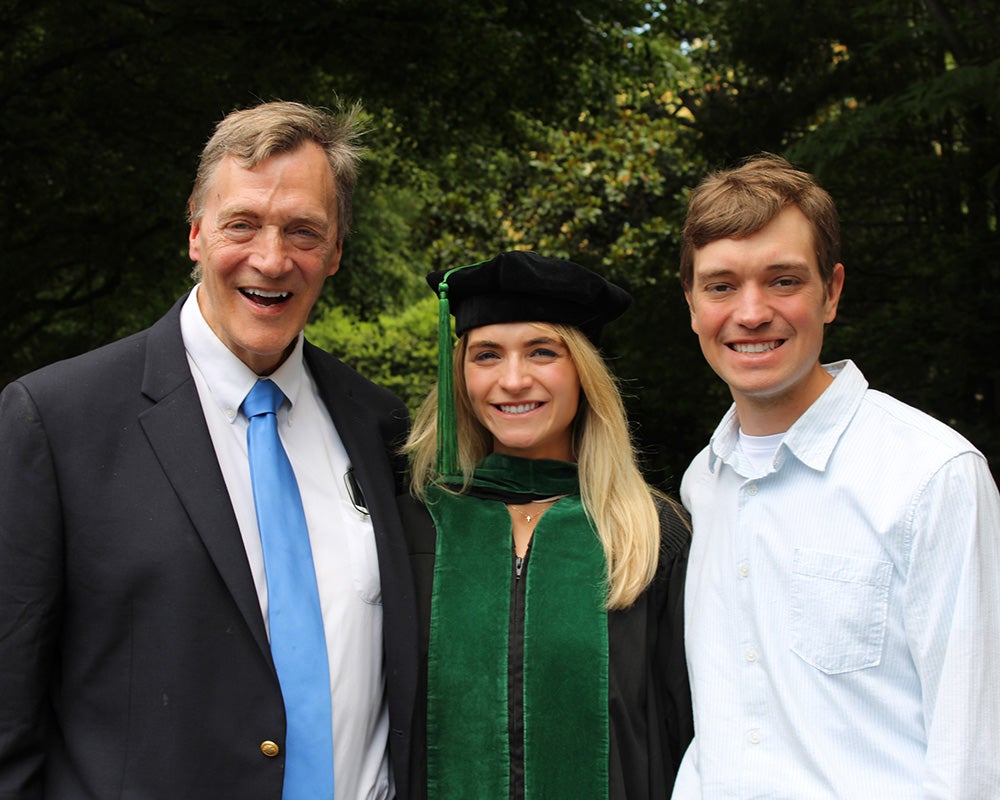 Carlynn Winters in academic regalia stands with her father and brother outdoors on campus