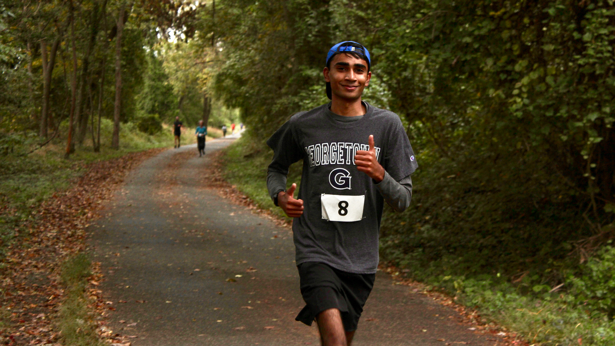 A runner on a paved trail through the woods gives two thumbs up