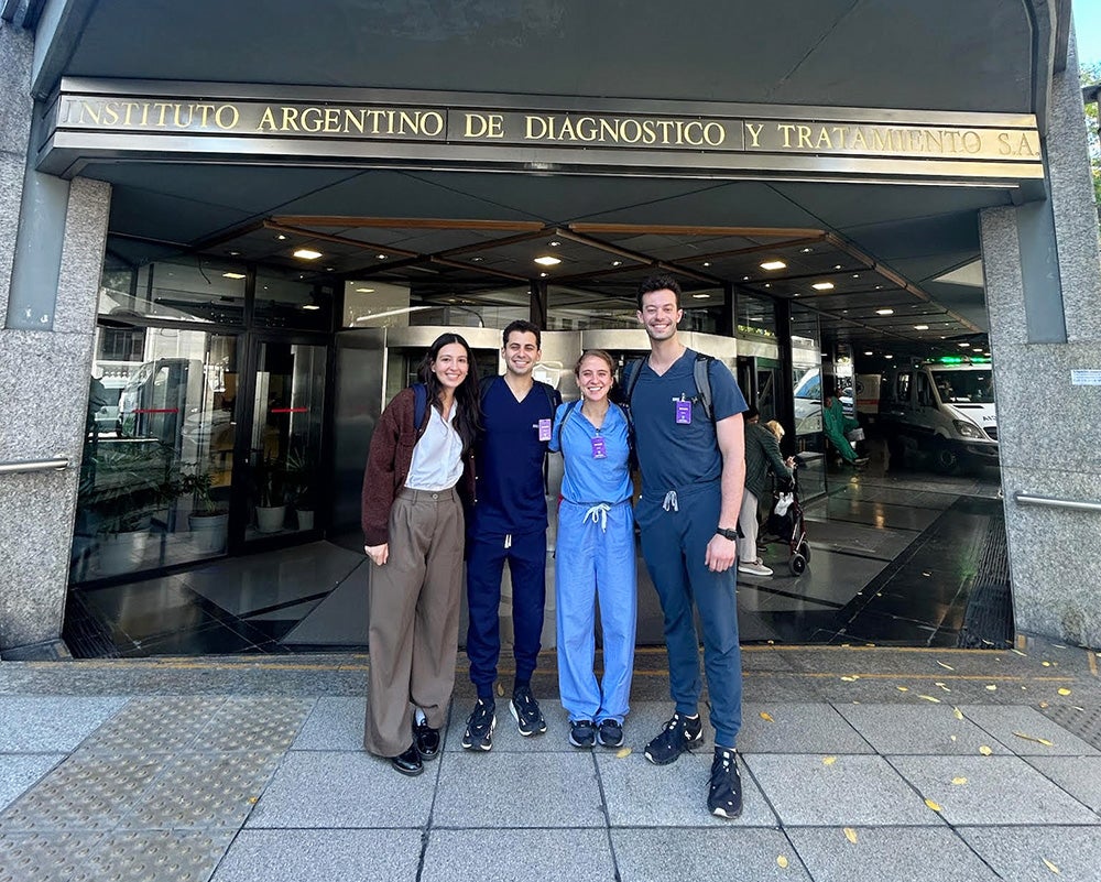 Four medical students stand together in front of the door of a building with the name on the marquee above the door