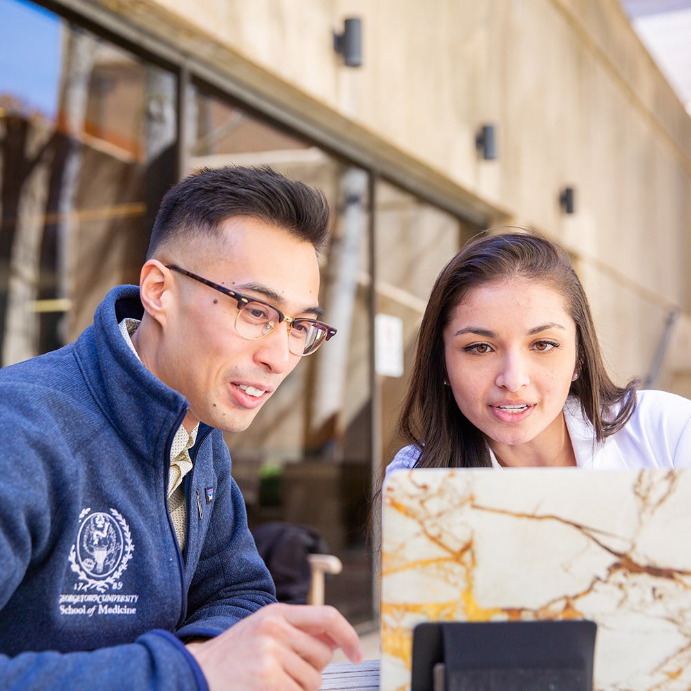 Two med students look at a computer screen while seated at an outdoor table