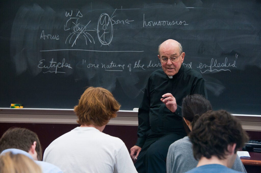 A priest sits at the front of a class with a chalkboard behind him