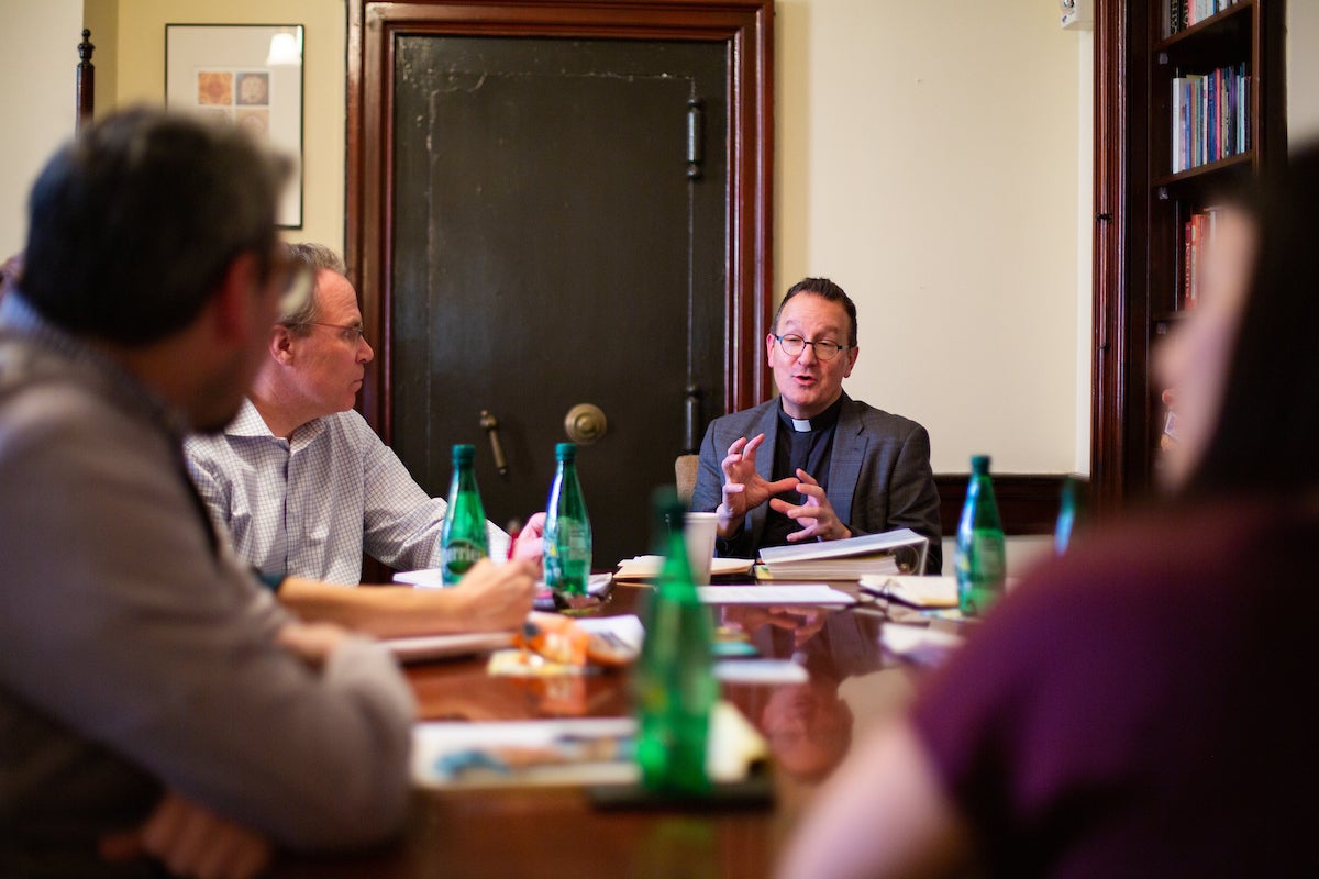 A Jesuit priest speaks while seated at a table with other scholars