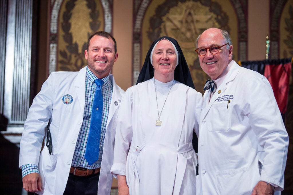 A medical student stands with a nun and Professor Mitchell at the White Coat Ceremony