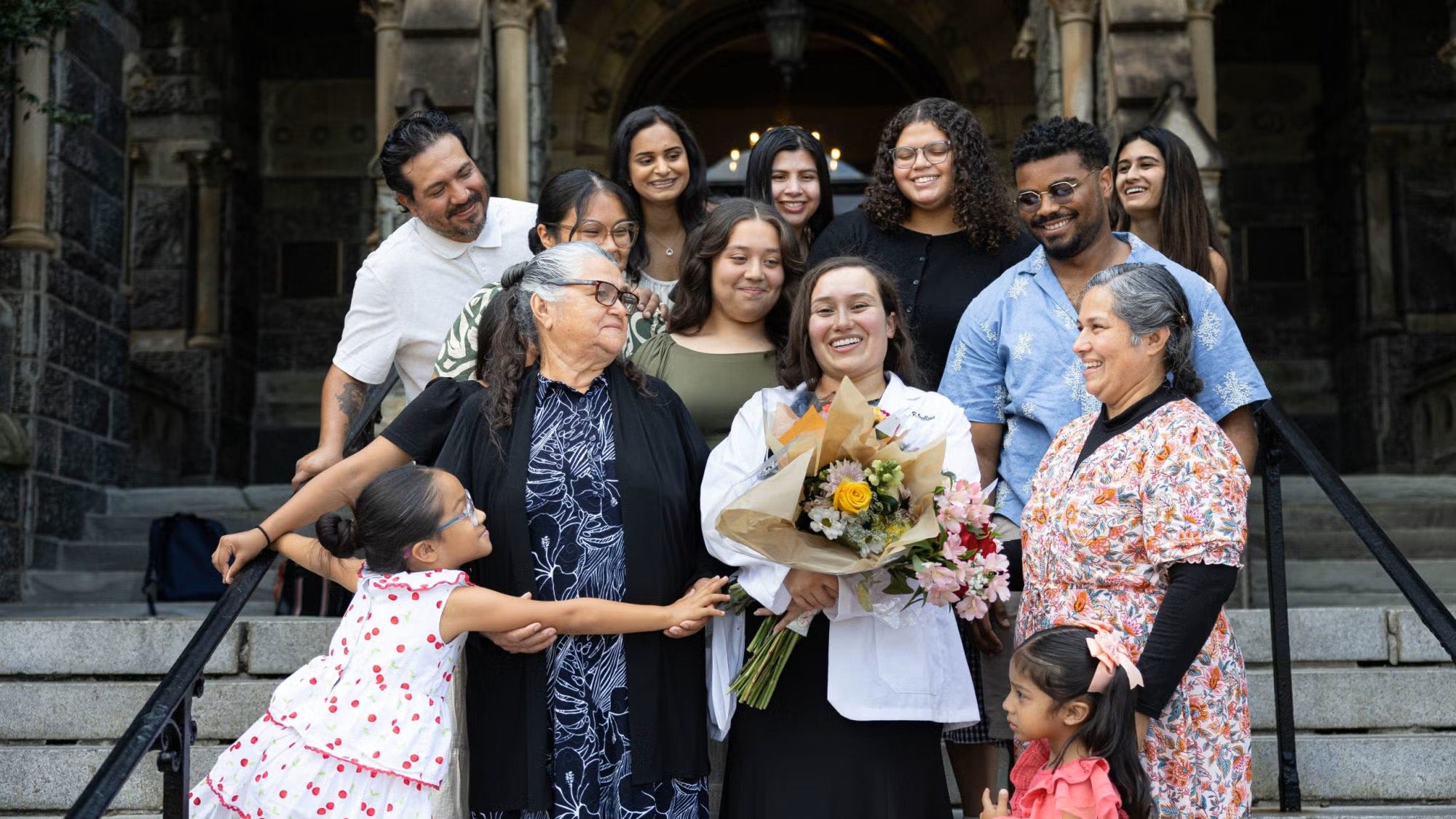 Halyn Orellana wears a white medical coat and is surrounded by her family members as they all stand on the steps of Healy Hall