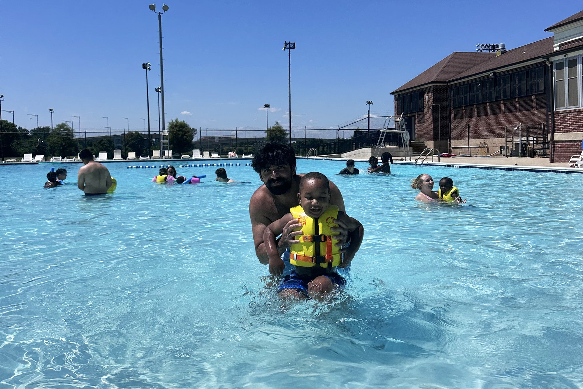 A med student holds a young boy wearing a lifejacket in a pool; other swimmers are in the background