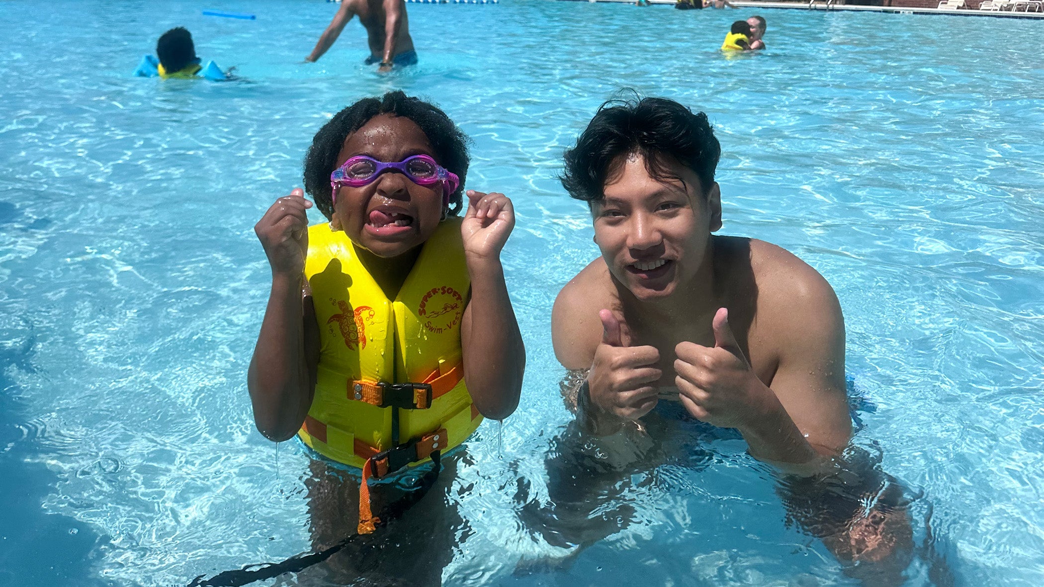 A medical student gives two thumbs up next to a camper making a funny face while swimming in a pool