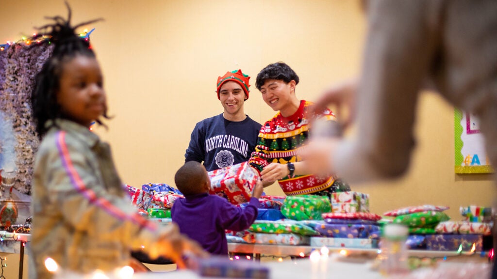 Two medical students hand a wrapped present to a boy while a girl in the foreground also receives a gift