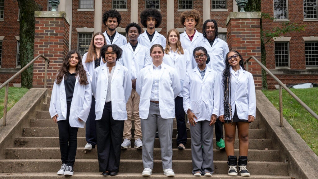 A group of students wearing medical white coats stand together on the steps in front of the Med-Dent Building on campus