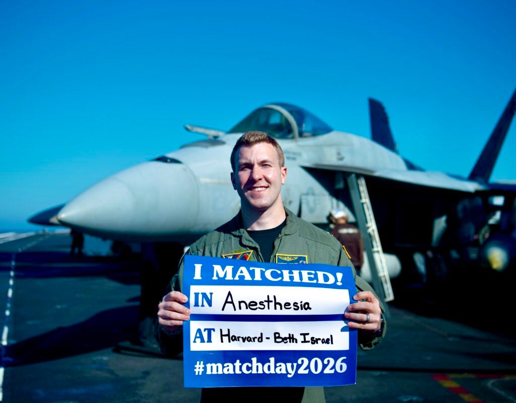 Dr. Matthew Civilette, class of 2022, standing on board the flight deck of the USS Nimiz, holding up a sign with his 2026 residency match.  