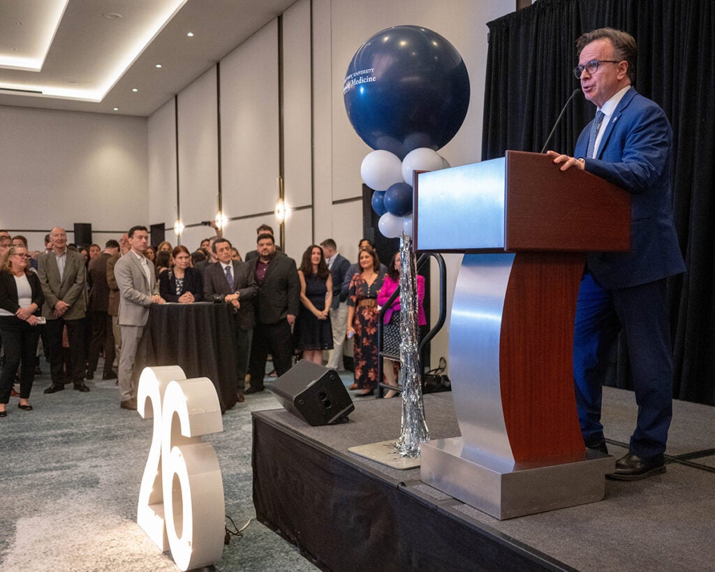 Dr. Beauchamp speaks from a podium on a dais set up at the front of the room on Match Day while students and families look on