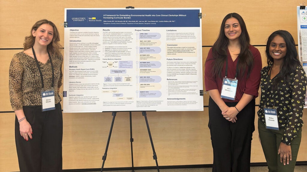 Three female students stand near a poster on an easel