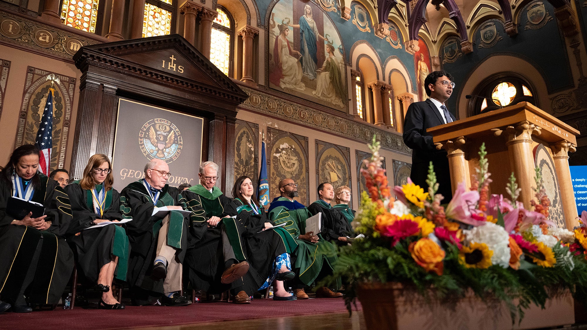 Arjun Mathur speaks from a podium onstage in Warwick Evans Auditorium with School of Medicine faculty seated behind him
