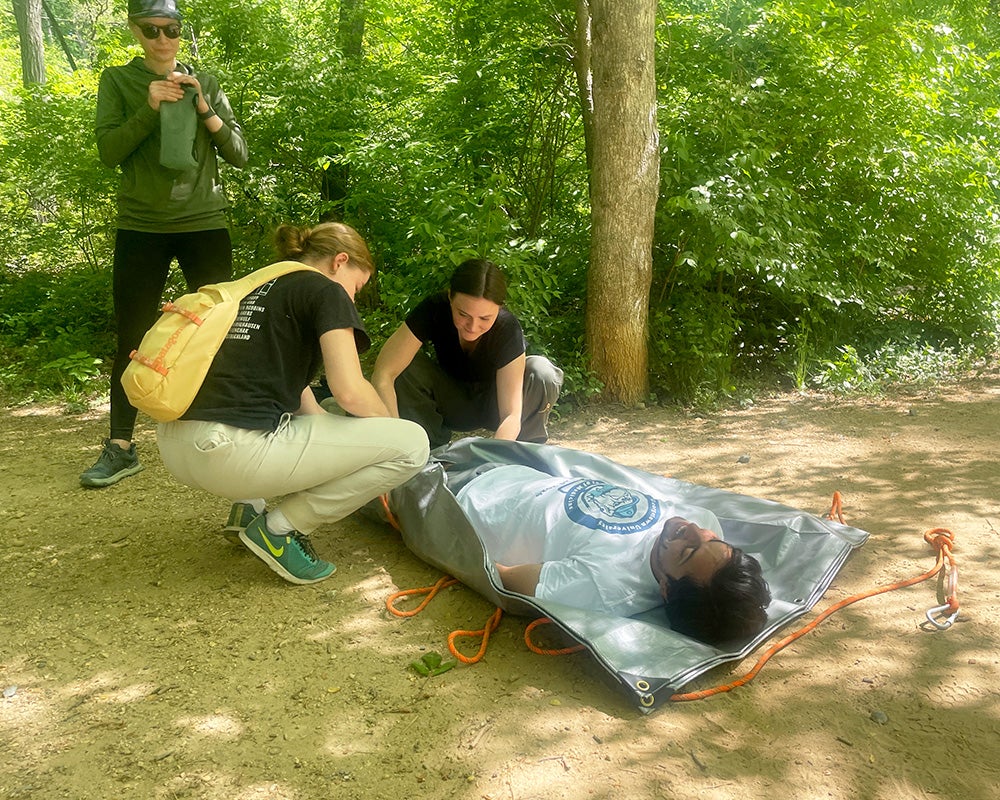 Three Wilderness Medicine students create a stretcher for an individual lying prone during a mock rescue