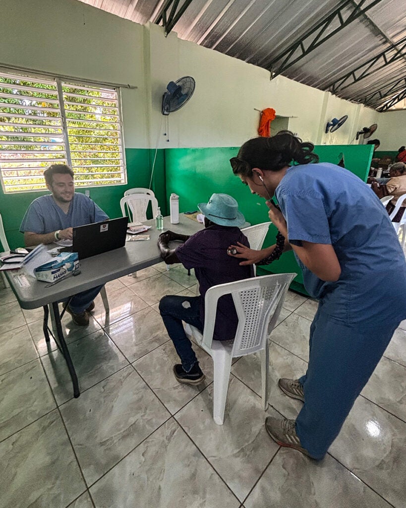 A medical student listens to a man's heartbeat with a stethoscope while another records notes on a laptop