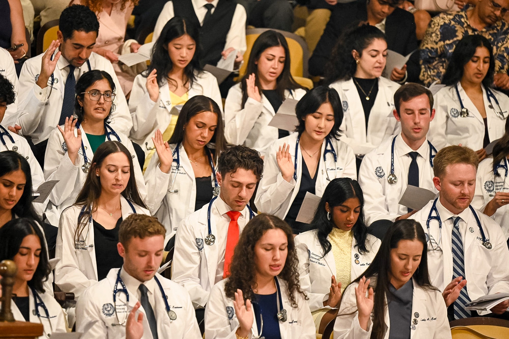 Rows of medical students in white coats hold up their hands as they recite the Hippocratic Oath