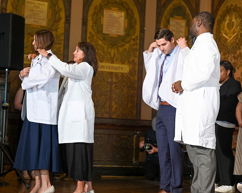 Two faculty members each help a student put on a white medical coat
