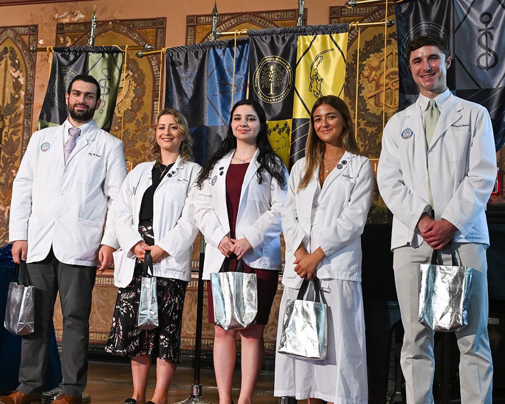 Five medical students in white coats holding silver bags stand together