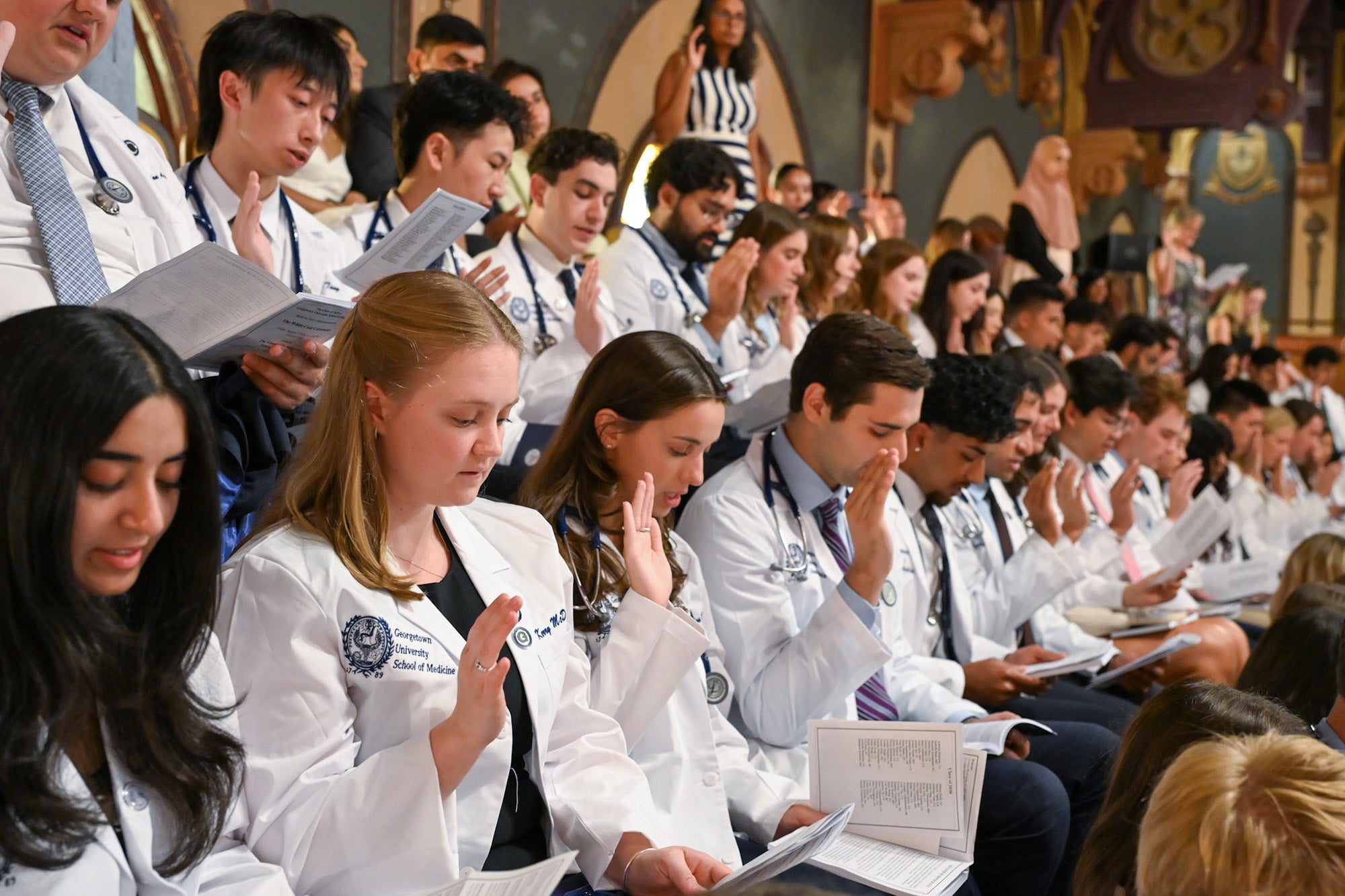 Rows of medical students wearing white coats hold their hands up and recite the Hippocratic Oath